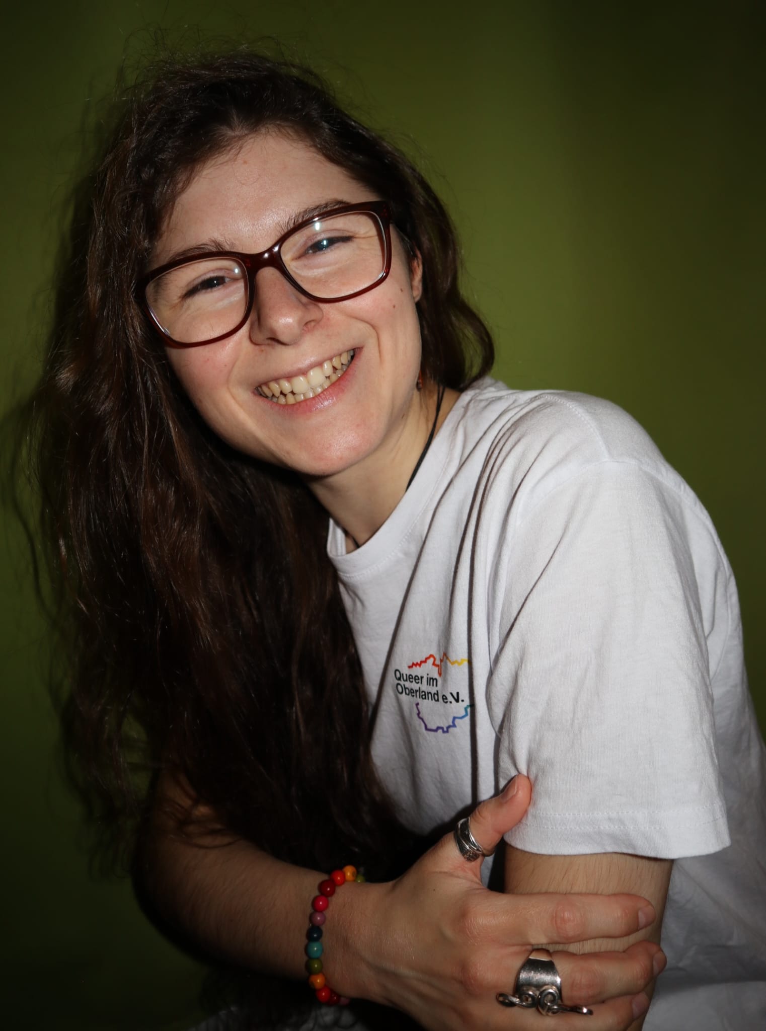 Die Person auf dem Foto trägt ein weißes Shirt mit aufgedrucktem Logo des Vereins Queer im Oberland e.V. und lächelt in die Kamera. Zudem trägt die Person eine braune Brille, ein wenig Schmuck sowie lange, dunkelbraune, etwas lockige Haare, die offen über die Schulter fallen.
